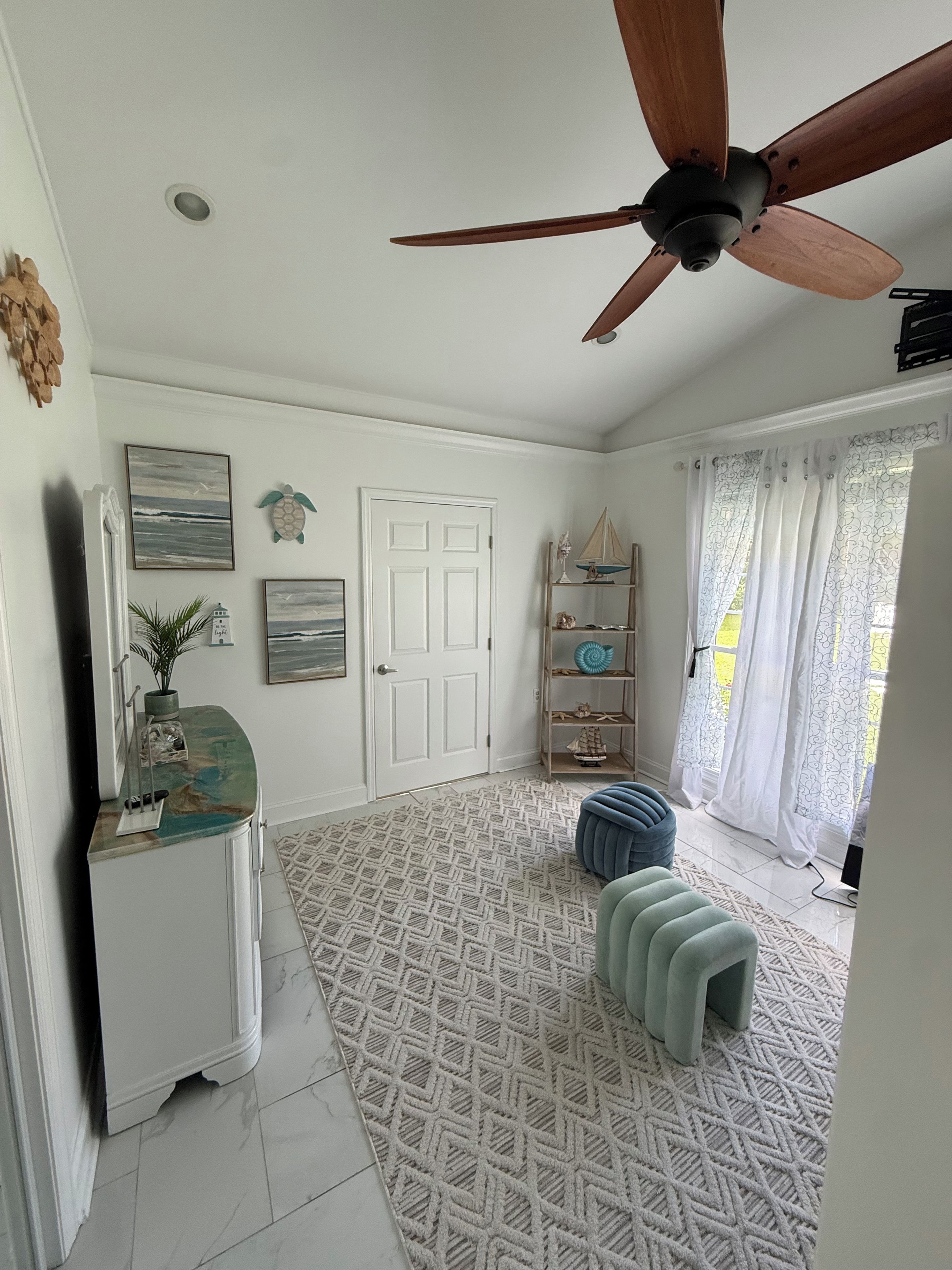 A room with a ceiling fan, a white door, a patterned rug, and a small blue ottoman.