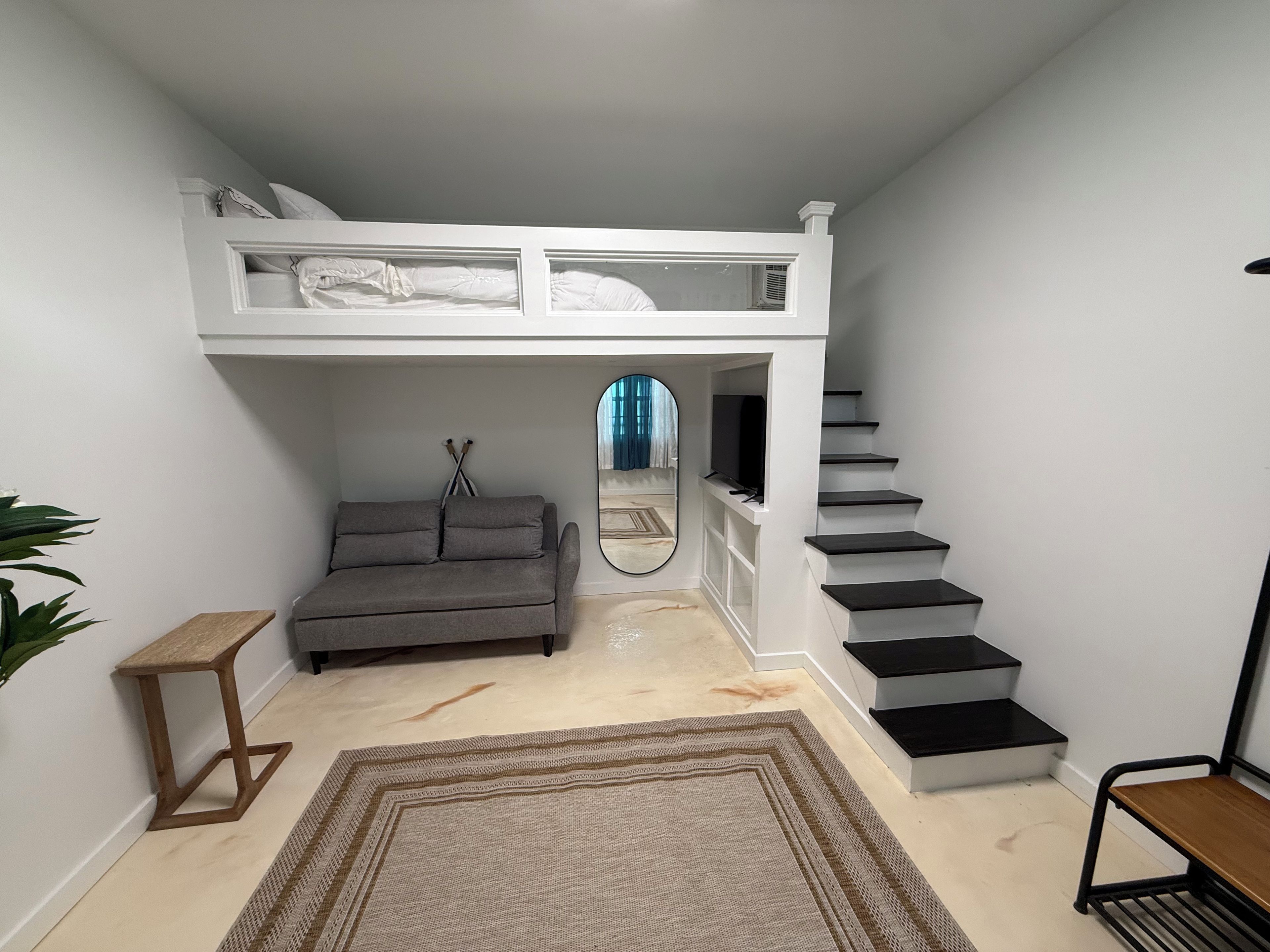 A loft bedroom with a bunk bed, a grey sofa, a small wooden table, a mirror, and a staircase.