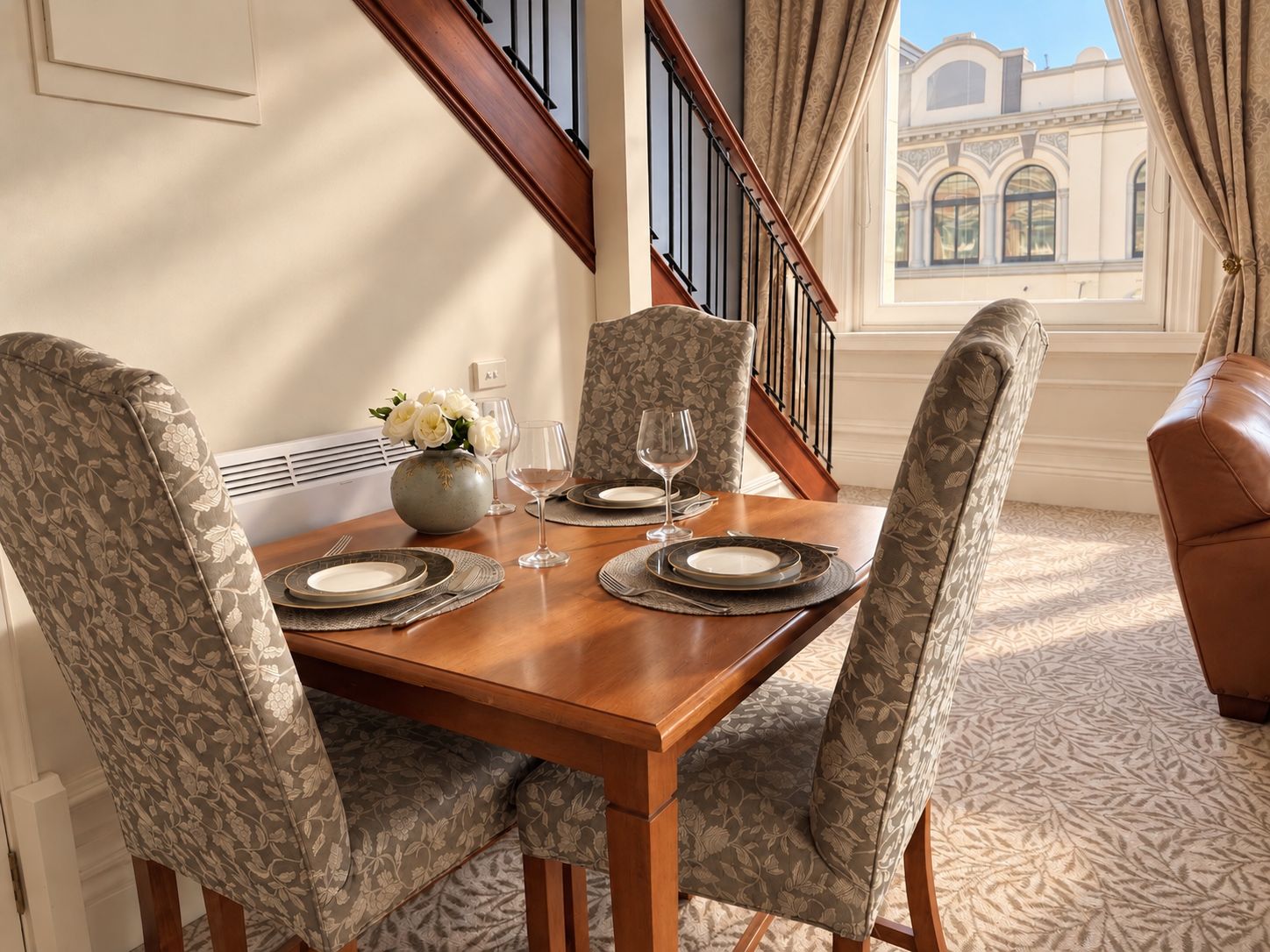 A dining area with a wooden table set for four, surrounded by patterned chairs, a vase with flowers, and a view of a building through a window.