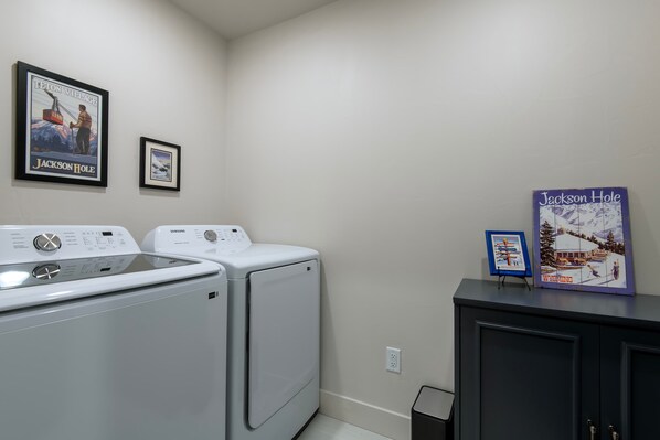 A laundry area with two front-loading washers and dryers, a black cabinet, and two framed posters on the wall. - Teton Homestead: Stone Peak Hideaway (Driggs)