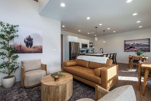 A modern living room with a brown sofa, a wooden coffee table, and a dining area with wooden chairs and a table. - Teton Homestead: Stone Peak Hideaway (Driggs)