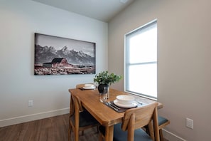 A dining area with a wooden table, chairs, a vase with plants, and a painting of a mountain landscape on the wall. - Teton Homestead: Stone Peak Hideaway (Driggs)
