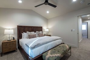 Un dormitorio con cama, mesitas de noche, ventilador de techo y una puerta que da a otra habitación.