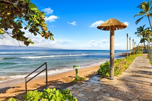 A beach with a stone pathway, a caution sign, and a thatched umbrella. - Kaanapali Sunset Escape with Ocean Views (Lahaina)