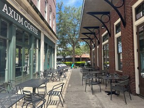 An outdoor cafe area with tables and chairs, a brick building with a sign reading 'AXUM COFFEE', and a clear sky. - Winter Garden Butterfly Retreat - Trailside (Winter Garden)