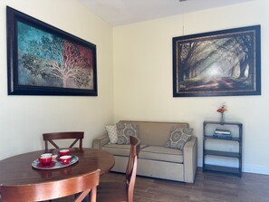 A dining area with a round table, chairs, and a coffee setup. There is a sofa and a bookshelf with books and a vase of flowers. Two framed paintings are on the walls. - Winter Garden Butterfly Retreat - Trailside (Winter Garden)