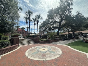 A courtyard with a patterned pavement, brick walls, palm trees, and a building with a red roof in the background. - Winter Garden Butterfly Retreat - Trailside (Winter Garden)