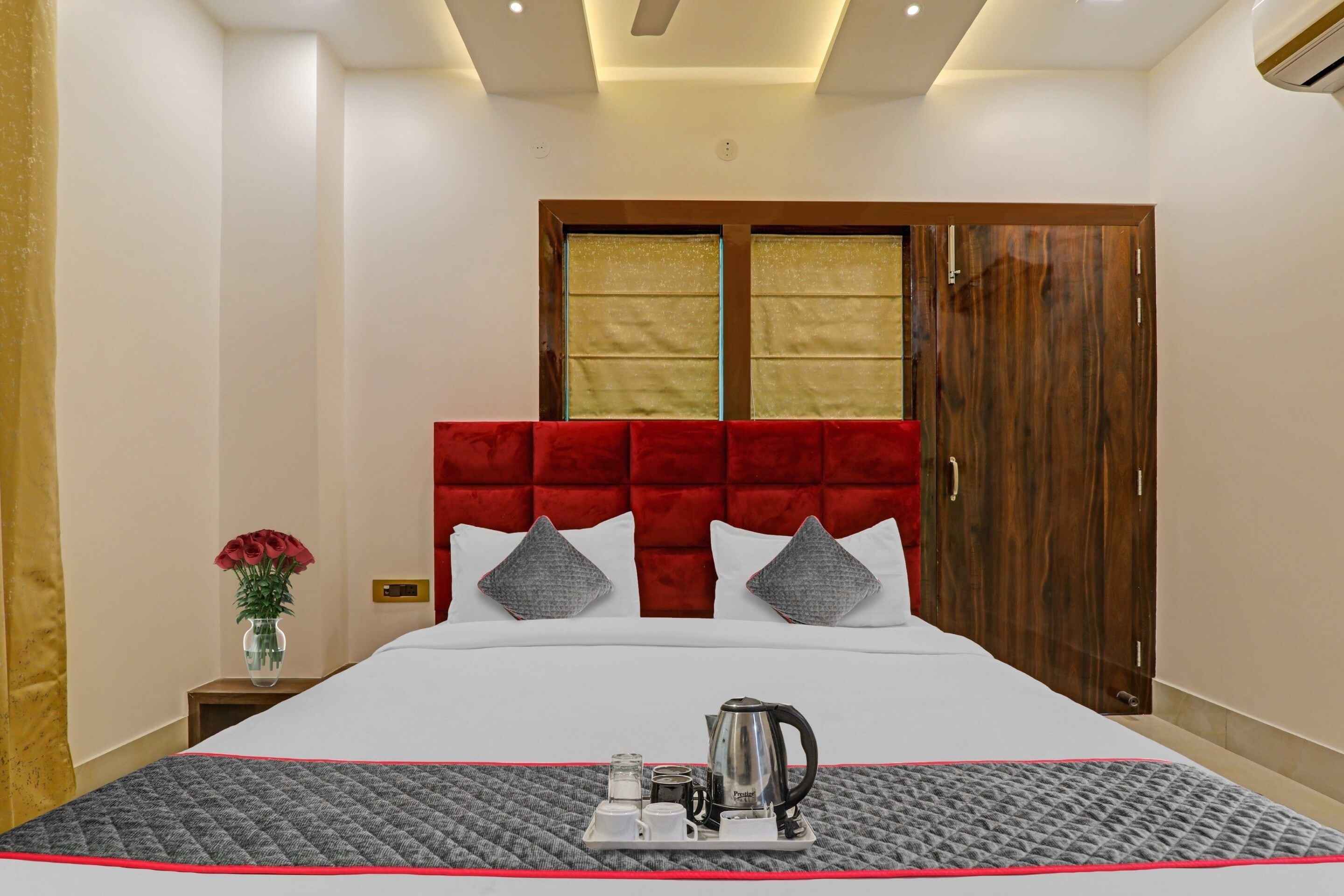 A hotel room with a bed, a bedside table with a vase of flowers, a tray with a kettle and cups, and a wardrobe with a mirror.