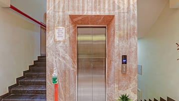 An elevator with a digital display, a sign requesting social distancing, and a staircase with a hand sanitizer dispenser.