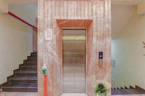An elevator with a digital display, a sign requesting social distancing, and a staircase with a hand sanitizer dispenser.