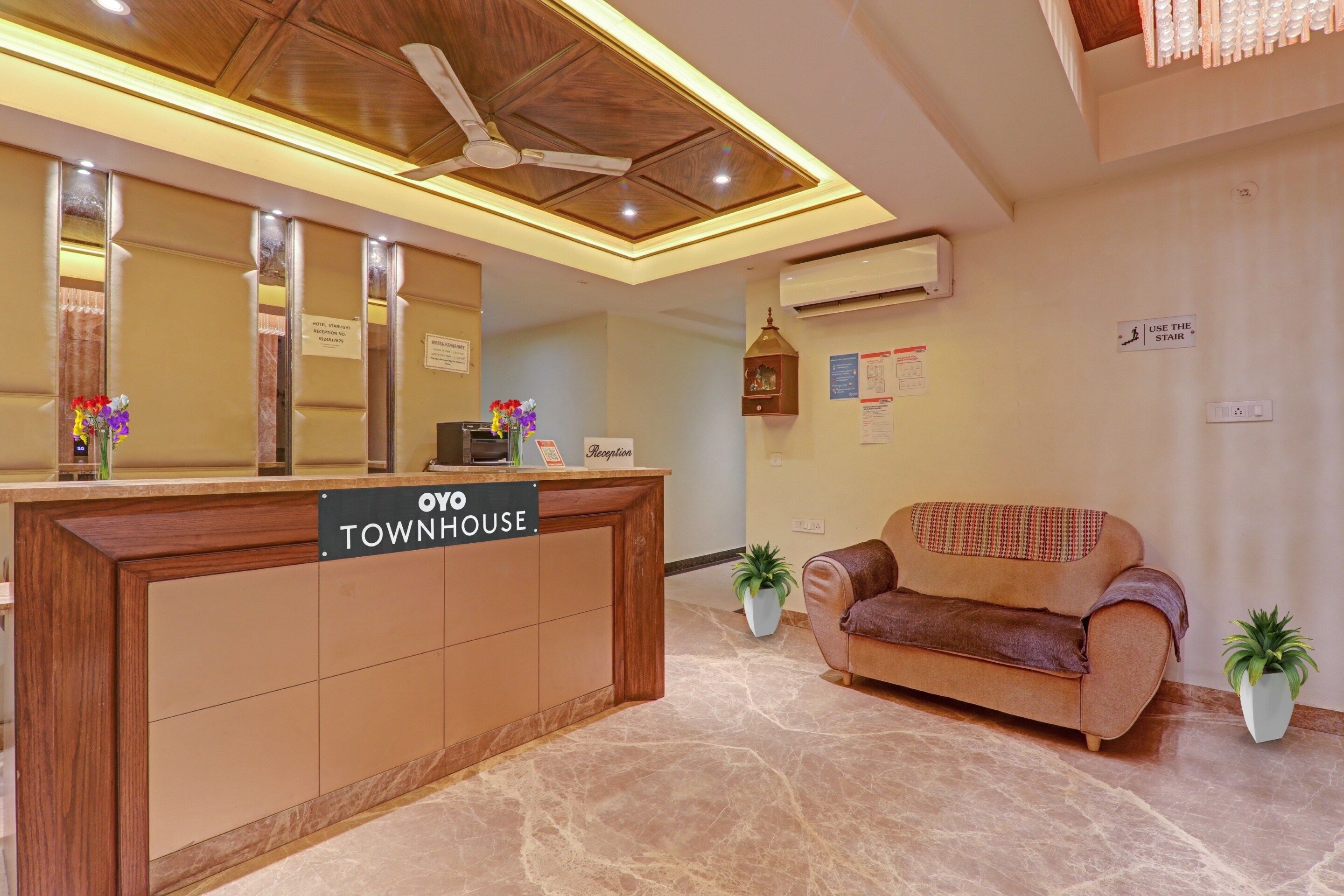 A reception desk labeled 'OYO TOWNHOUSE' with a ceiling fan, air conditioning unit, and a reception area with a couch and potted plants.