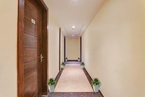A hallway with a wooden door on the left, numbered 202, leading to a room. The hallway has a tiled floor and is flanked by walls with potted plants.