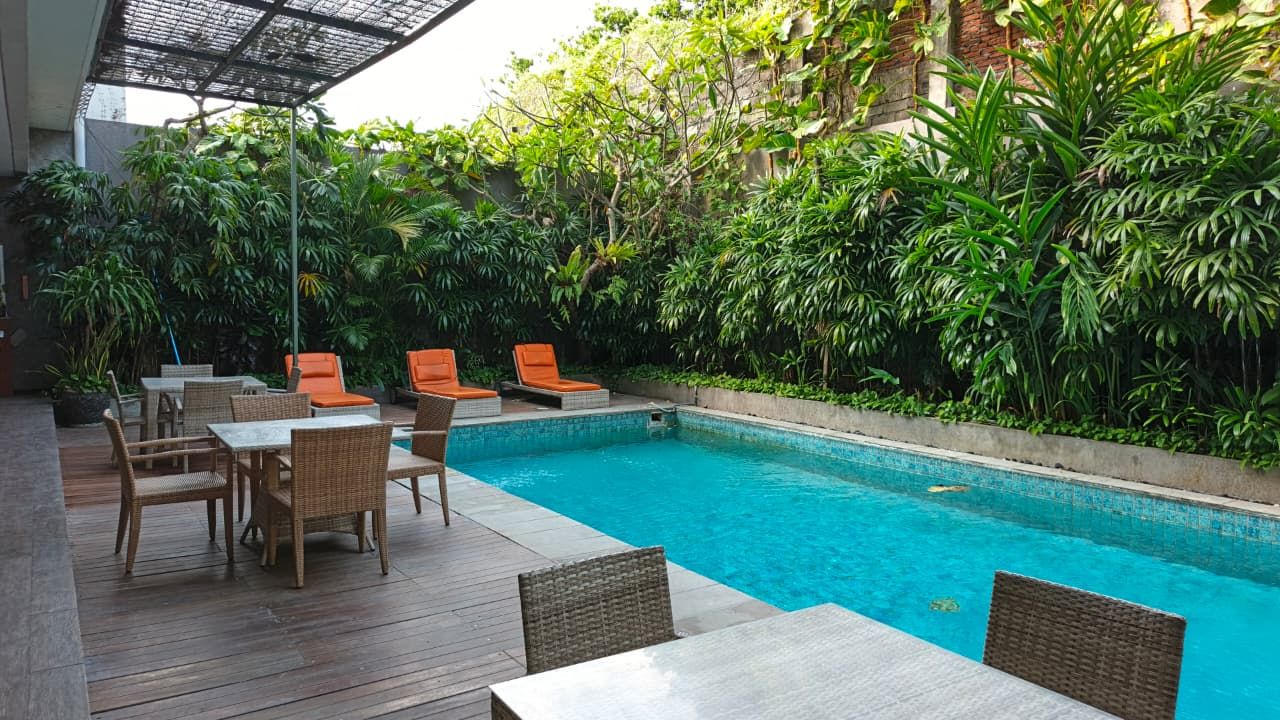 Zona de piscina con mobiliario de mimbre y sillas naranjas, rodeada de abundante vegetación.