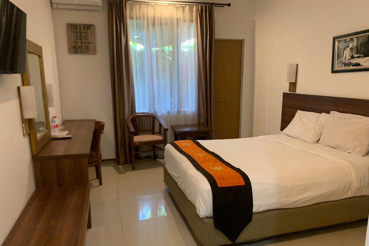 A hotel room with a bed, a desk, a chair, a window with curtains, and a door.