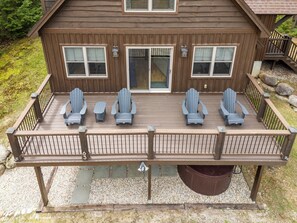 Eine Holzterrasse mit vier Adirondack-Stühlen, einem kleinen Tisch, einem überdachten Patio und einer Steinsäule im Hintergrund.