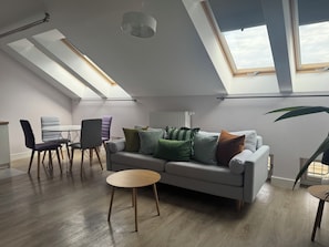 A modern living space with a high ceiling, skylights, a grey sofa, a dining table with chairs, and a potted plant. - Barbican Apartments by Fragola (Kraków)