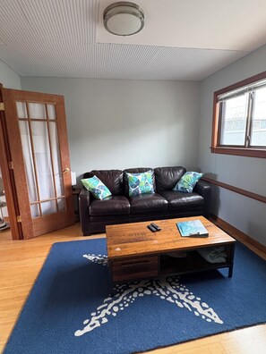 A room with a brown leather sofa, a wooden coffee table, a blue patterned rug, a wooden door, and a window with blinds. - Sweet and Private Apartment Close to Everything Beautiful and Fun! (El Granada)
