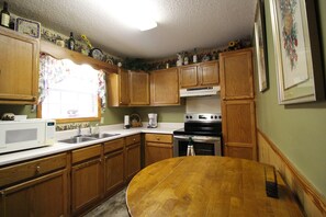 A kitchen with wooden cabinets, a microwave, a stove, and a dining table. - Bear Kottage Creekside Cabin (Bryson City)