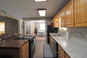 A kitchen with wooden cabinets, a central island with a stove, and a dining area with chairs. - Captain's Bend Creekside Cabin (Bryson City)