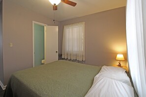 A bedroom with a bed, a ceiling fan, a window with curtains, a bedside lamp, and a door leading to another room. - Wolf's Den Creekside Cabin (Bryson City)