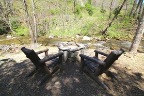 Ein rustikaler Außenbereich mit zwei Holzstühlen, die einem Steinkaminfeuer zugewandt sind, umgeben von einer bewaldeten Landschaft mit einem Bach.
