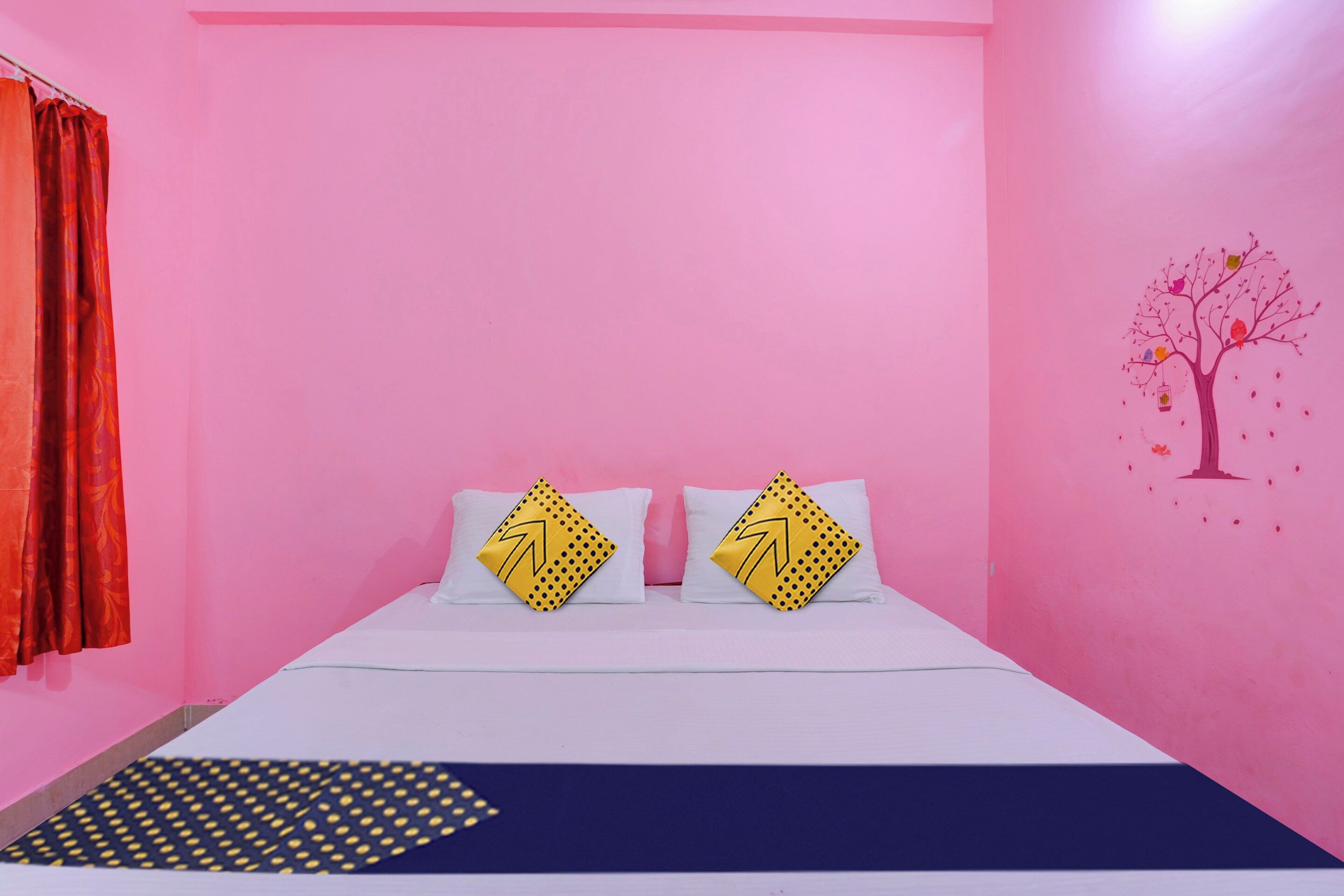 A bedroom with a pink wall, a bed with white bedding, two pillows with yellow patterns, and a tree mural on the wall.