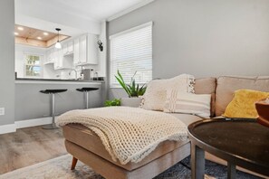A modern living room with a sofa, a coffee table, and a kitchen area with white cabinets and a bar stool. - The Quiet Oak - Peaceful South Tampa Condo w/ Pool and Reflection Pond (Tampa)