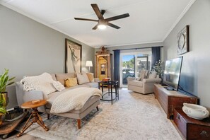 A living room with a sofa, armchairs, a coffee table, a TV, a ceiling fan, and a large window. - The Quiet Oak - Peaceful South Tampa Condo w/ Pool and Reflection Pond (Tampa)