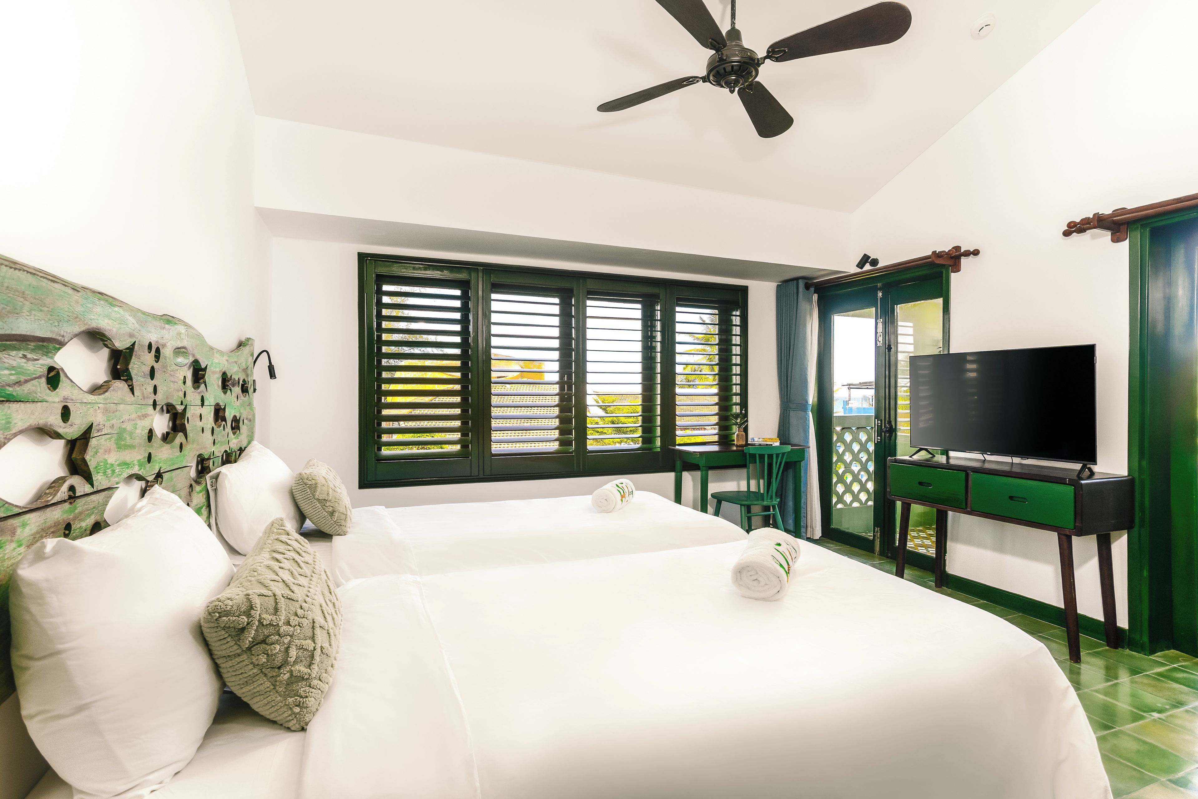 A hotel room with two beds, a ceiling fan, a TV on a stand, a window with shutters, and a green door.