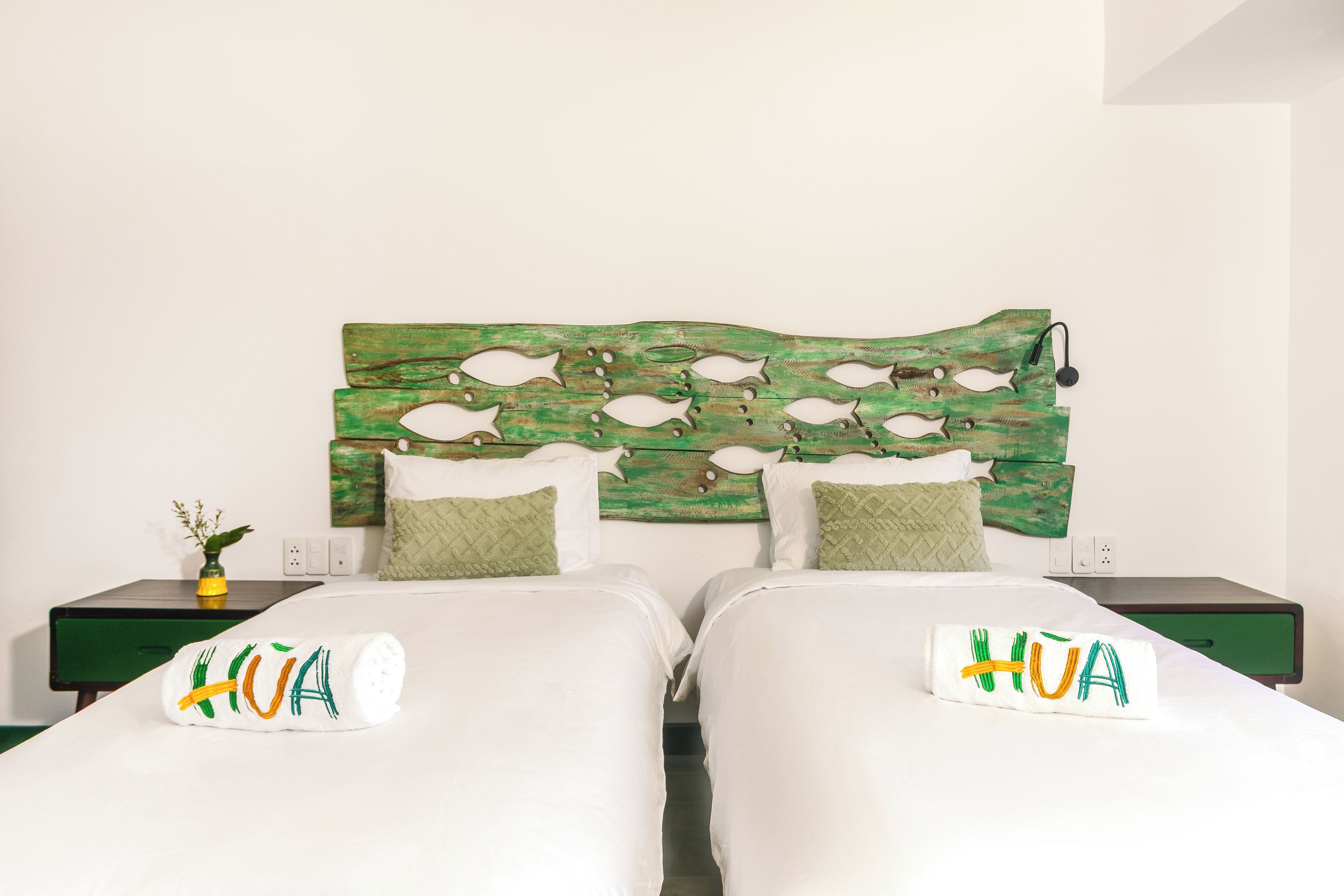 Cama de casal com cabeceira verde com desenhos de peixes, dois travesseiros e uma pequena mesa de cabeceira com uma planta.