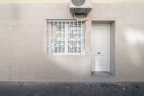 A building facade with a window featuring metal bars, a door with a small step, and an air conditioning unit mounted above the window. - GuestReady - Urban Studio in Barrio del Pilar (Madrid)