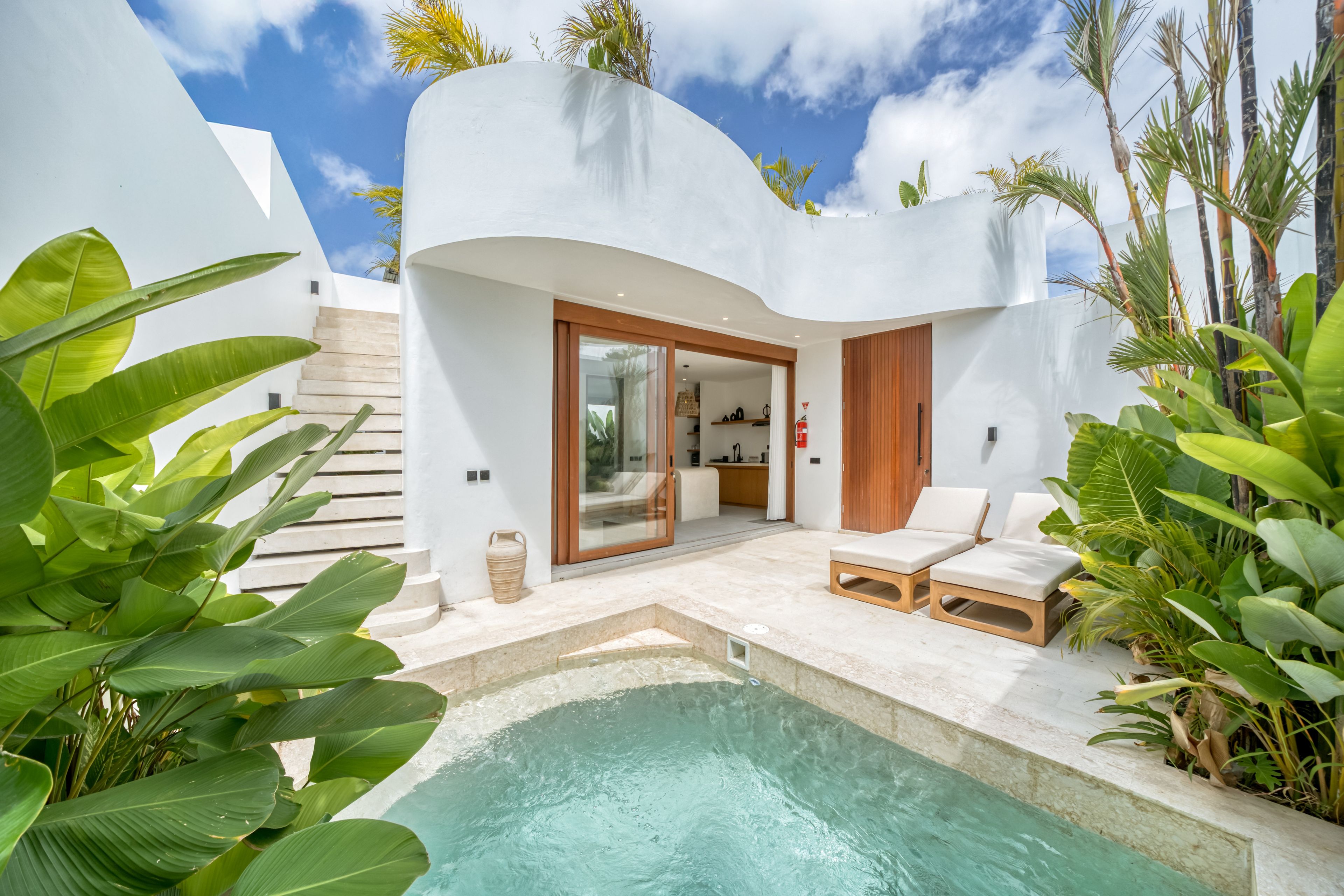 Un área de piscina moderna al aire libre con un edificio blanco curvo, una puerta de madera y un espacio de descanso con dos sillones de jardín.