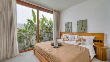 A modern bedroom with a large bed, a wooden headboard, and a view of tropical plants through a sliding glass door.