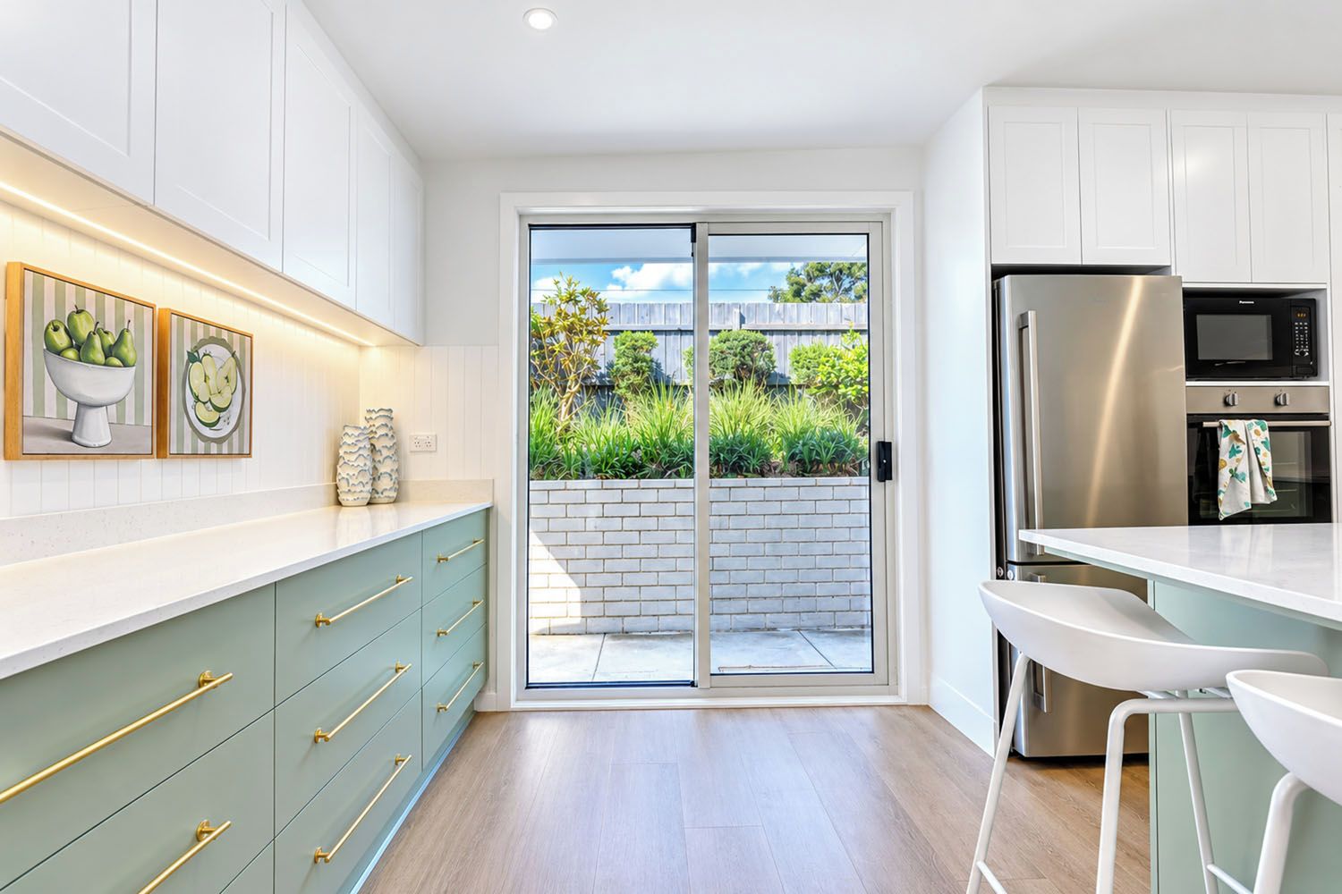A modern kitchen with white cabinets, a stainless steel refrigerator, and a sliding glass door leading to a garden.