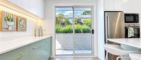 Una cocina moderna con armarios blancos, un refrigerador de acero inoxidable y un ventanal corredera que da a un jardín.