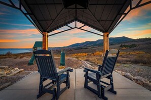 Twee Adirondack stoelen gericht op een speeltuin met een glijbaan, op een betonnen terras met uitzicht op een glooiend landschap en een wateroppervlak tijdens de zonsondergang.
