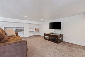 A living room with a brown leather sofa, a wooden coffee table, a flat-screen TV, and built-in shelving. - Summit Lakehouse -   Luxe Home, Sleeps 39, Game Room, Yard, Fire Pit! (Fish Haven)