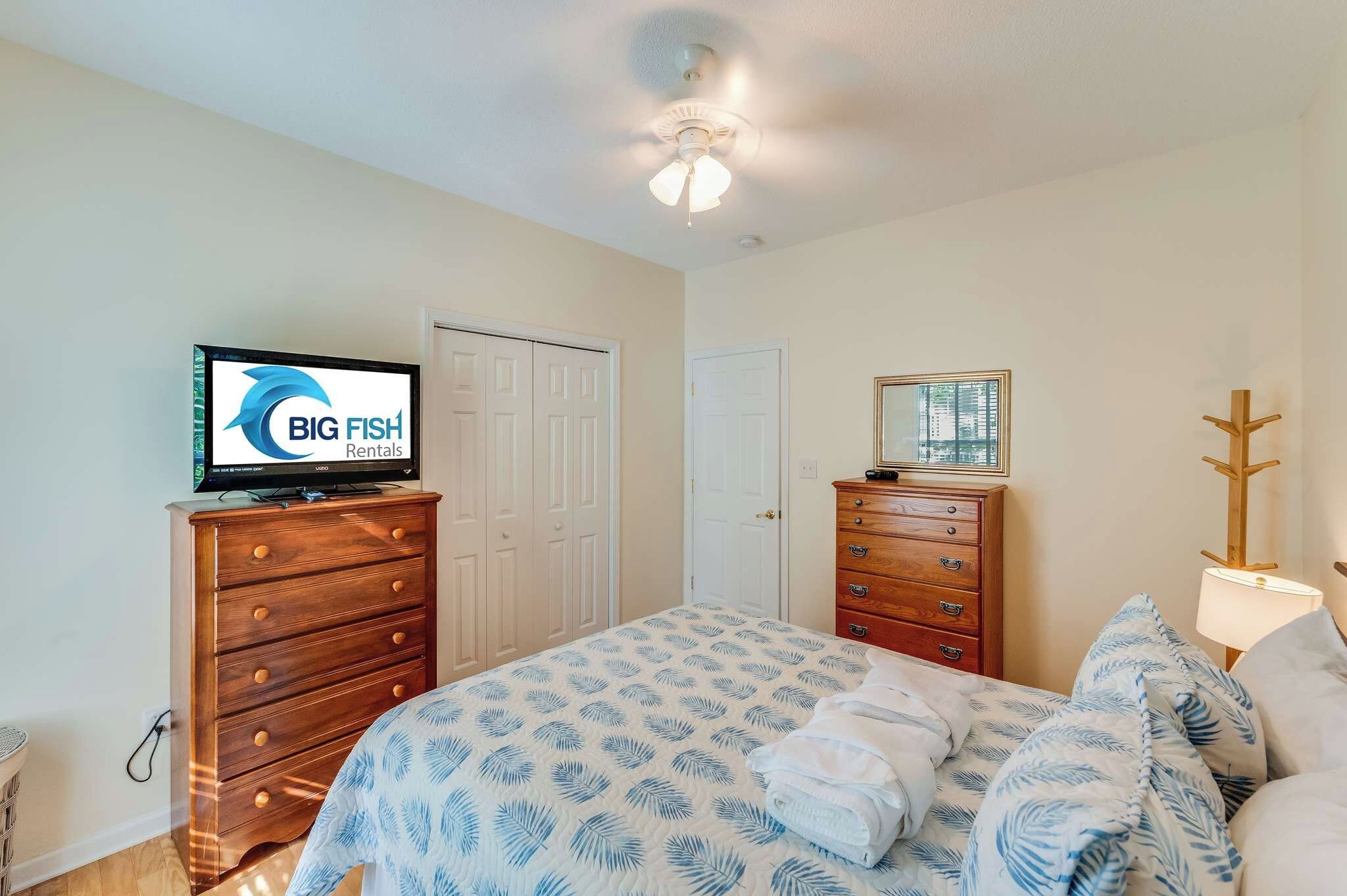 A bedroom with a bed, two wooden dressers, a television, and a ceiling fan.