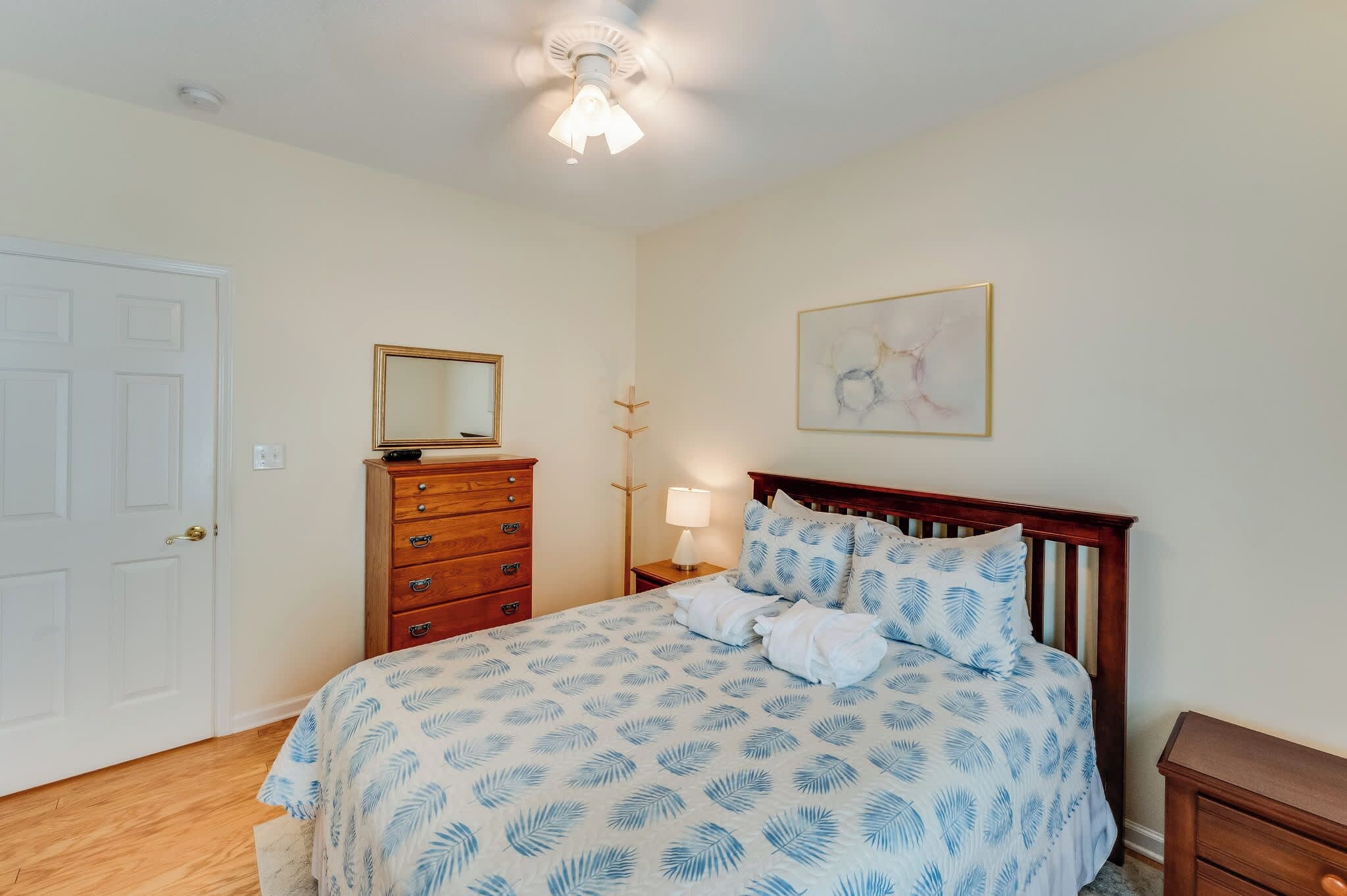 Une chambre à coucher comprenant un lit, une commode, une table de chevet, une lampe, un ventilateur de plafond, une porte et une œuvre d’art accrochée au mur.