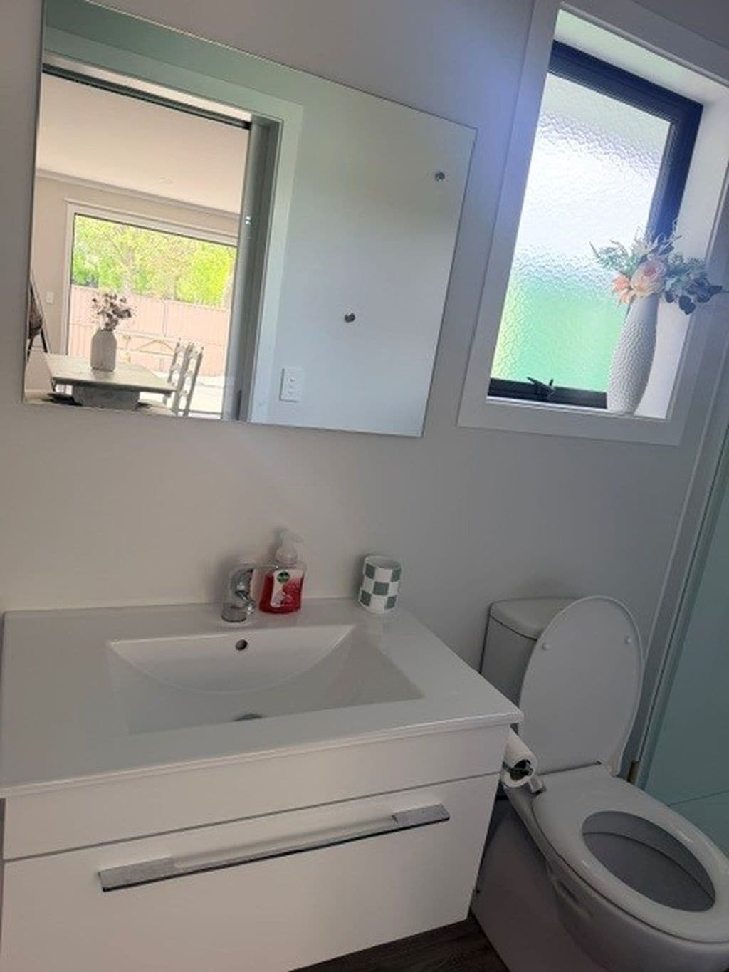Un baño con un lavamanos blanco, inodoro, espejo y ventana con vistas a la vegetación.