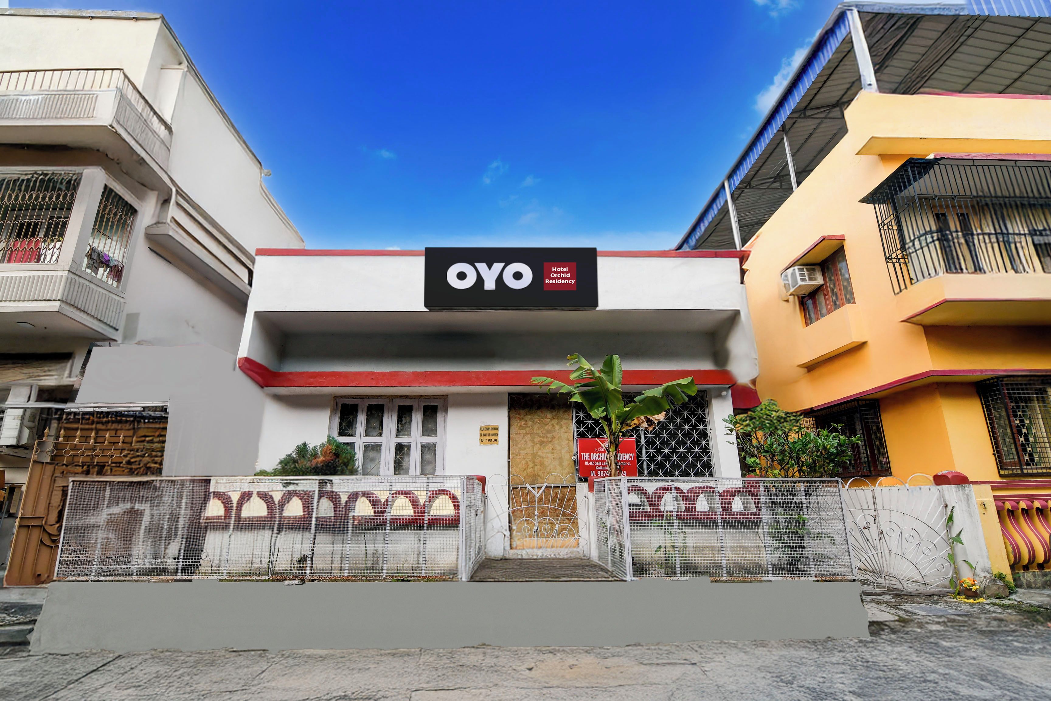 Un hotel llamado OYO con un letrero, una entrada protegida por una reja y un cielo azul despejado.