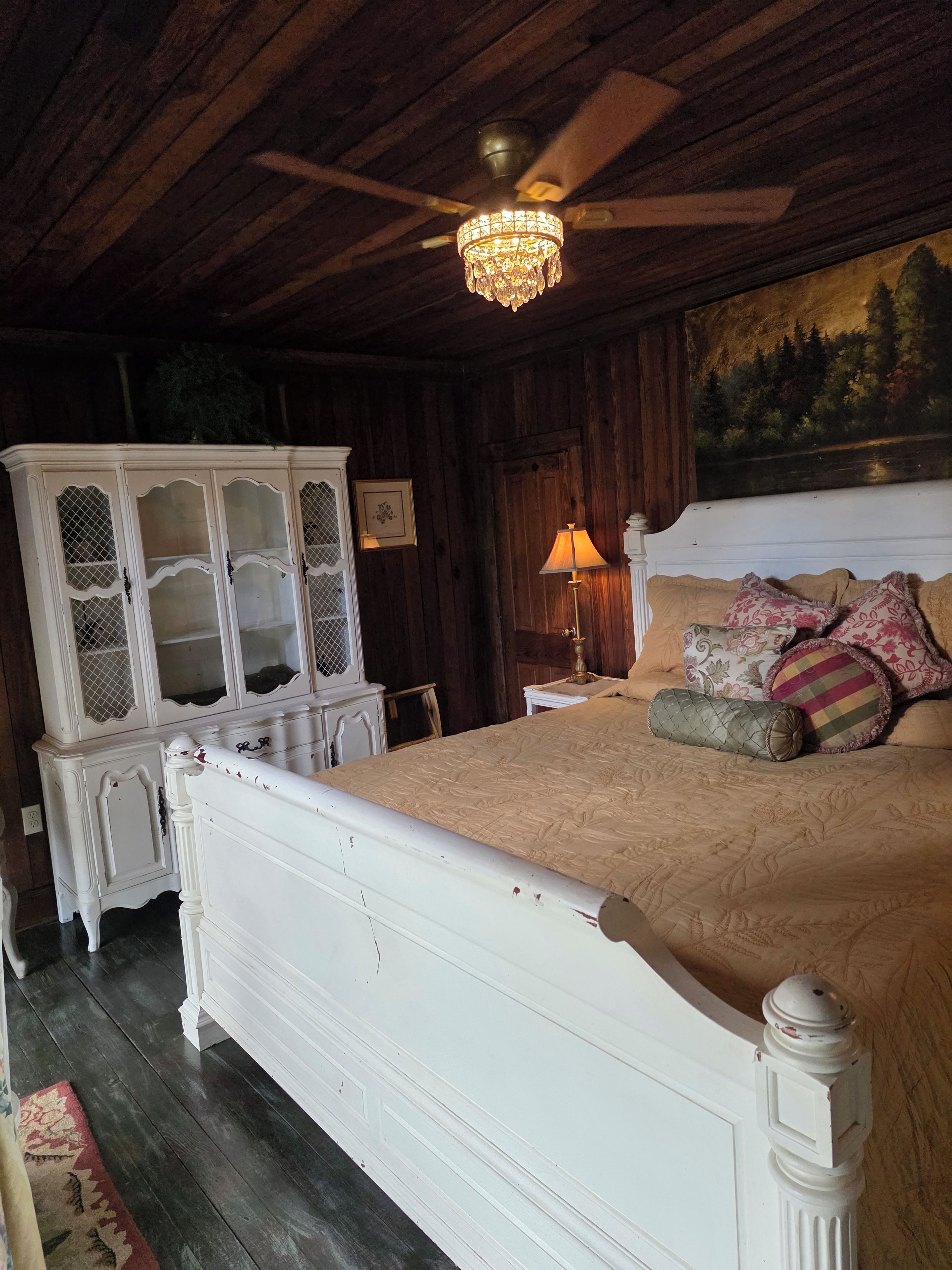 A bedroom with a white bed, a wooden ceiling, a chandelier, a painting, and a cabinet.