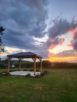 Um gazebo de madeira num campo gramado com um pôr do sol ao fundo.