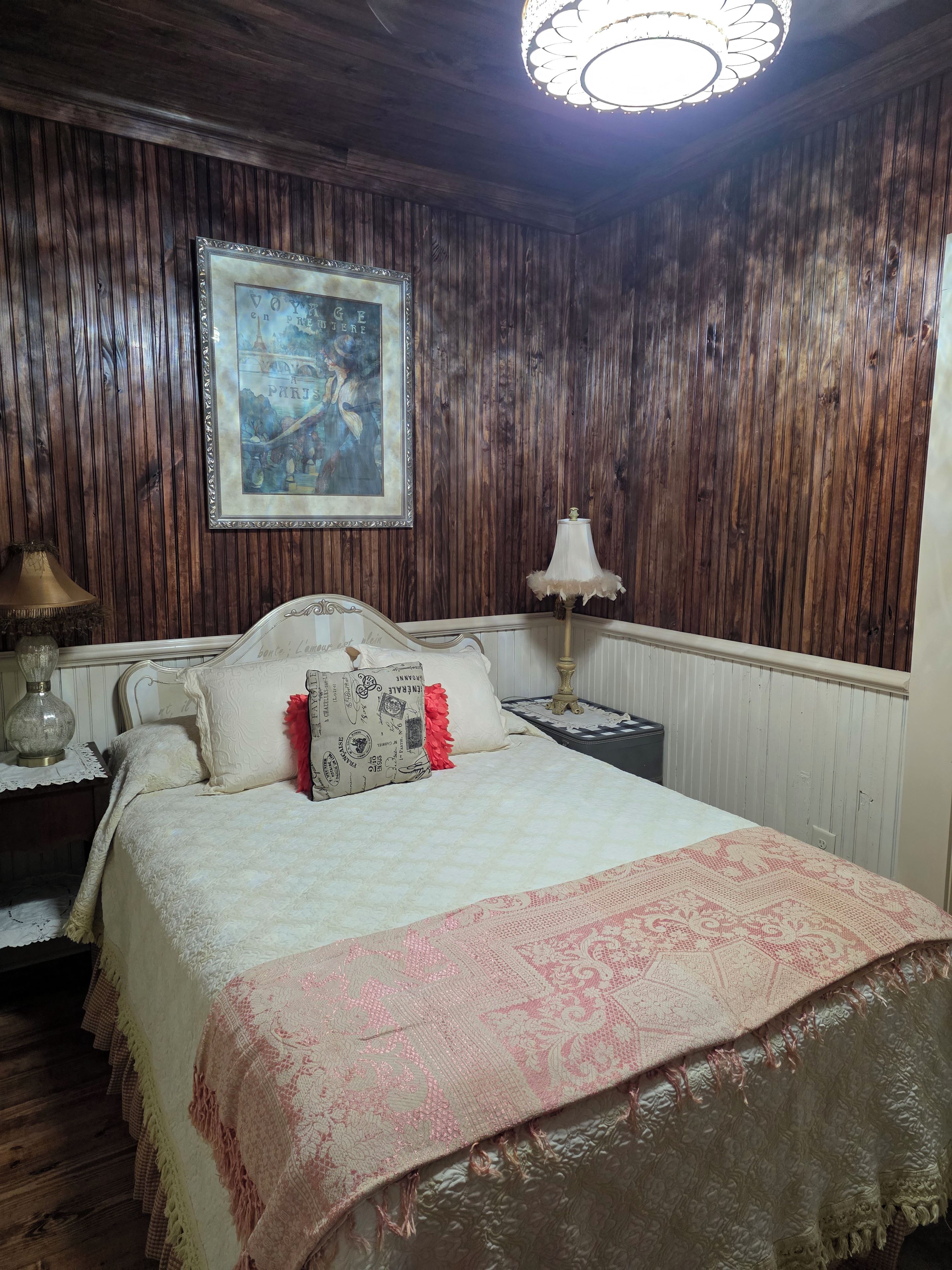 Quarto com parede revestida em madeira, cama com colcha estampada, quadro emoldurado e abajur na mesa de cabeceira.