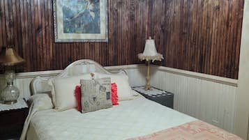 Quarto com parede revestida em madeira, cama com colcha estampada, quadro emoldurado e abajur na mesa de cabeceira.