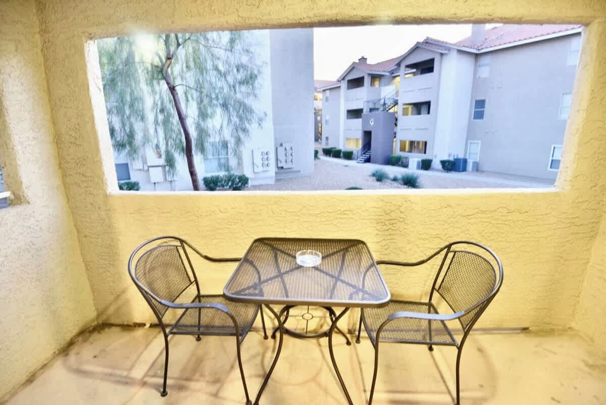 Een klein buitenterrein met een tafel en twee stoelen, een raam met uitzicht op een gebouw, en een gestructureerde muur.
