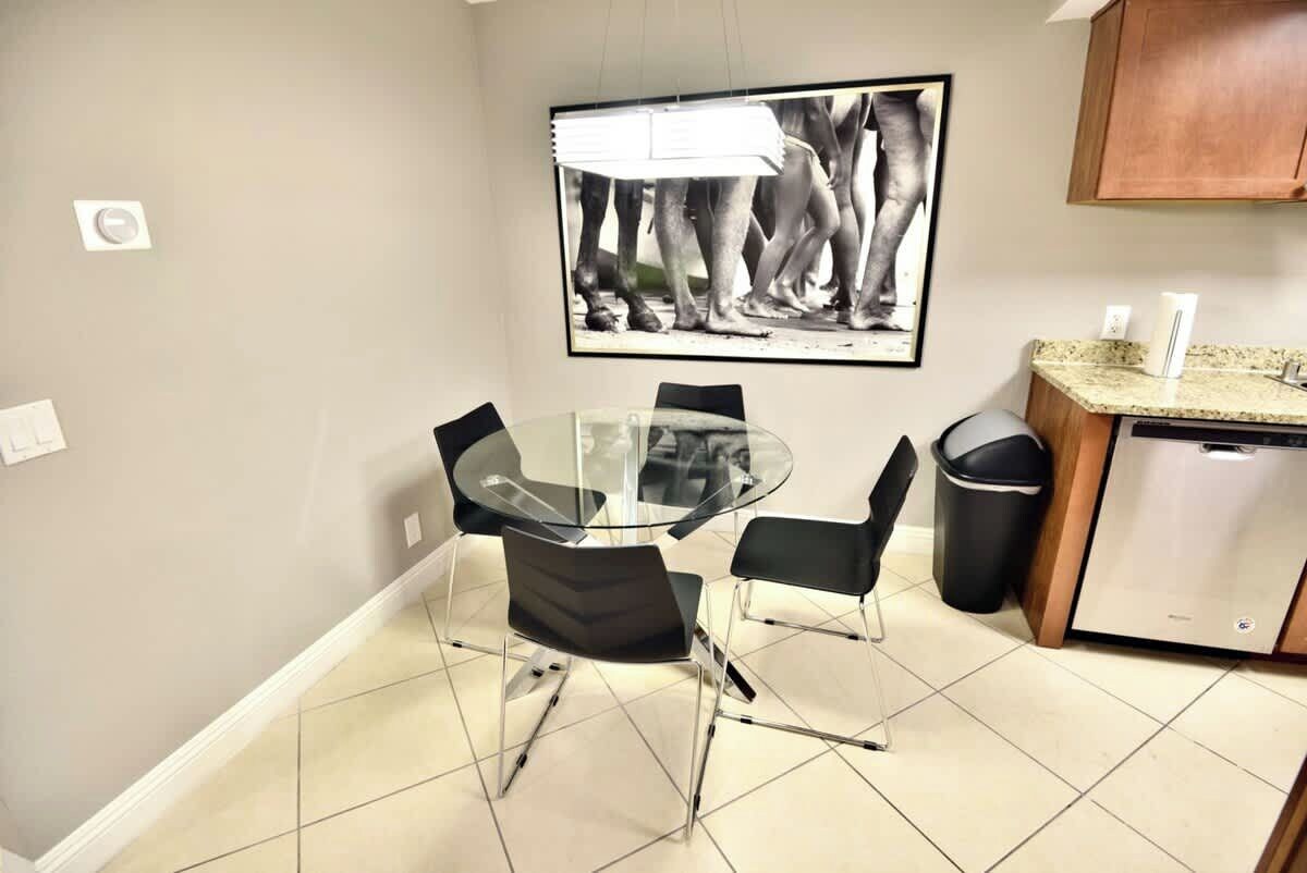 A small dining area with a glass table and four chairs, a black trash bin, a dishwasher, and a framed black and white photograph on the wall.