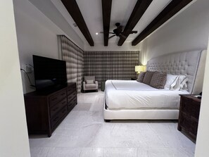 Un dormitorio con una cama grande, televisión, una silla y un ventilador de techo.