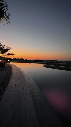 A calm waterfront scene with a paved walkway, palm trees, and a sunset.