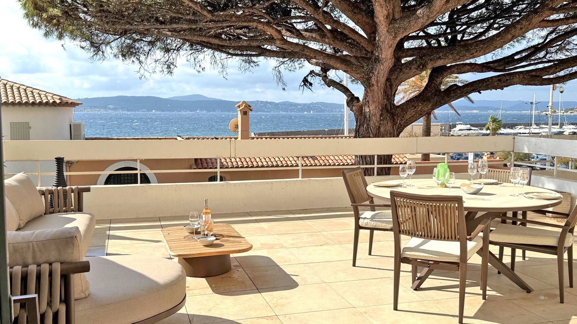 A terrace with a table and chairs, a large tree, and a view of the sea and boats.